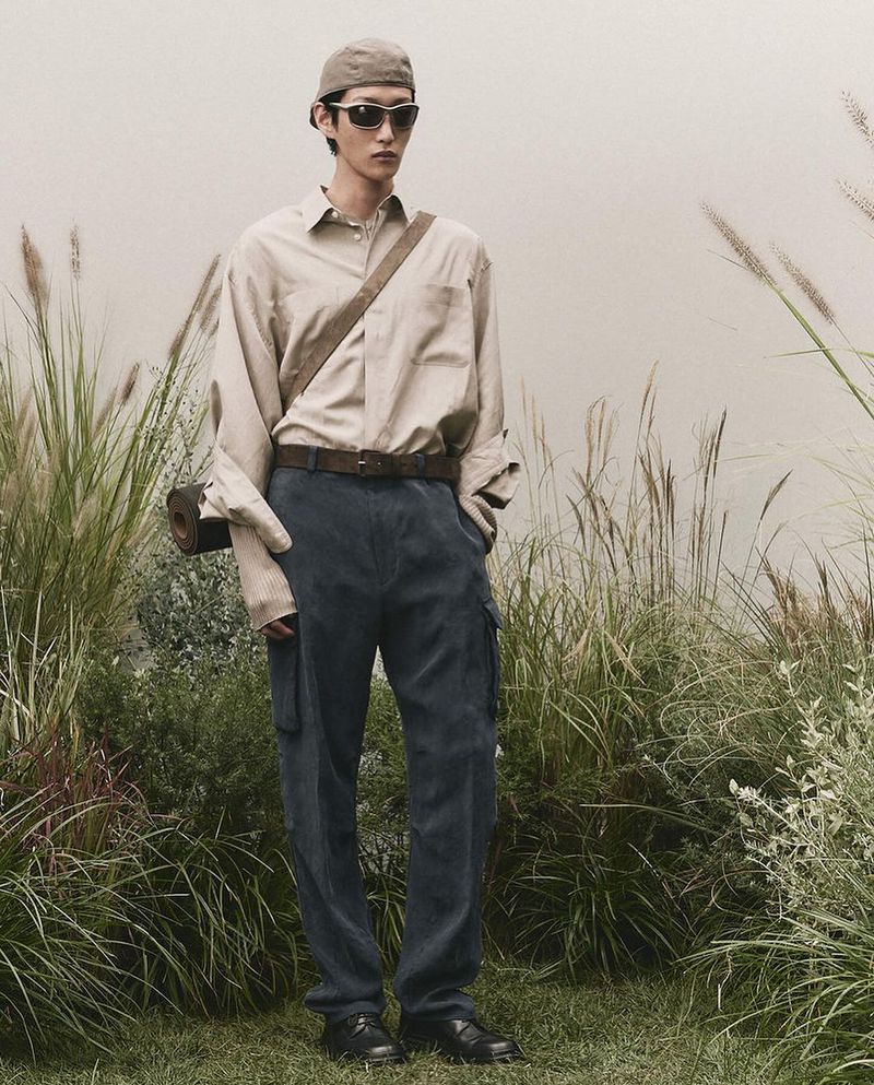 Full-length shot of a male model in an outdoor setting wearing a button-down shirt, trousers, sunglasses, and a cap, with grass in the background.