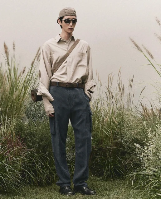 Full-length shot of a male model in an outdoor setting wearing a button-down shirt, trousers, sunglasses, and a cap, with grass in the background.
