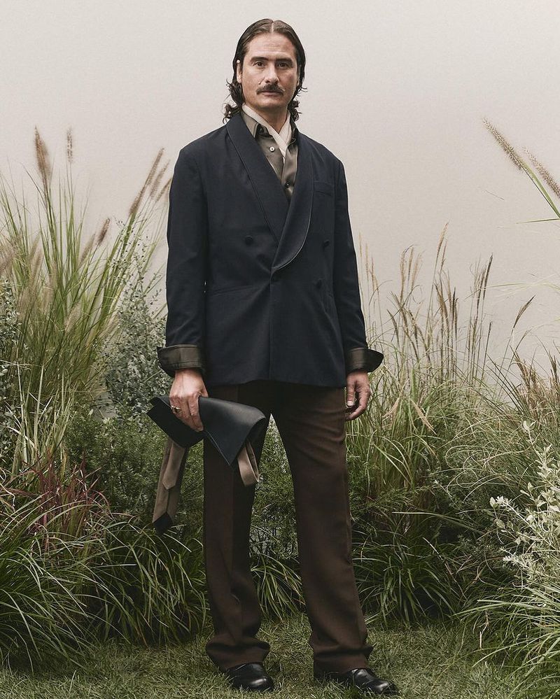Man wearing a dark double-breasted blazer and brown trousers standing amidst tall grass and plants