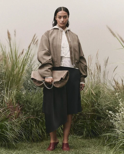 Adult female fashion model poses in front of grasses, wearing a jacket, skirt, shirt, handbag, and heels in an outdoor setting.
