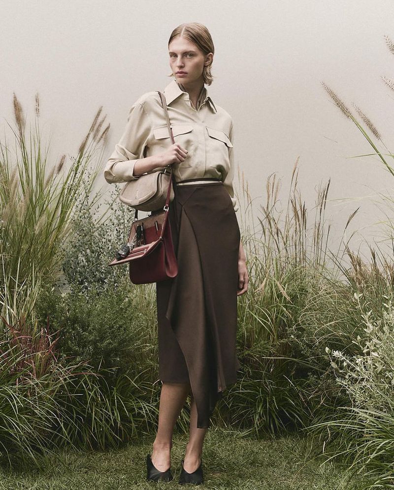 Adult female stands in a field of tall grass wearing a long brown skirt, light brown blouse, black high heels and carrying a brown purse.