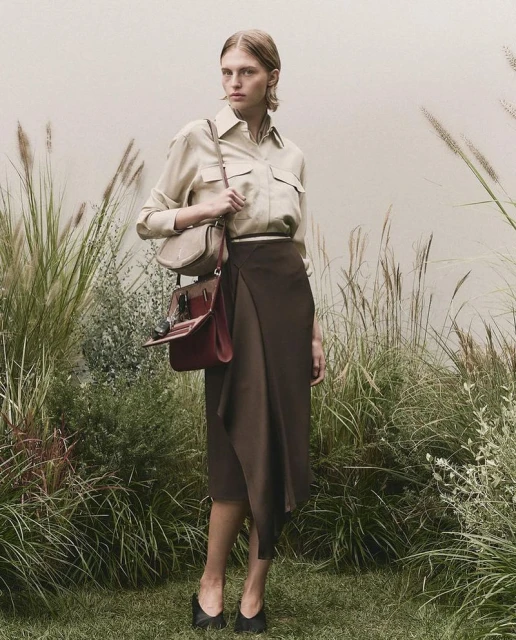 Adult female stands in a field of tall grass wearing a long brown skirt, light brown blouse, black high heels and carrying a brown purse.