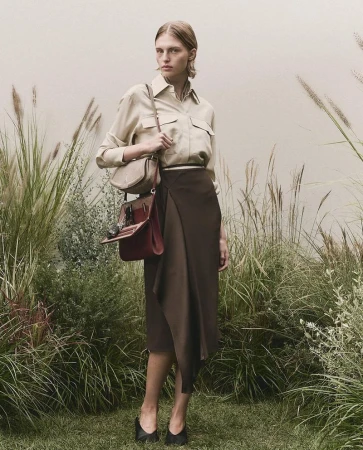 Adult female stands in a field of tall grass wearing a long brown skirt, light brown blouse, black high heels and carrying a brown purse.