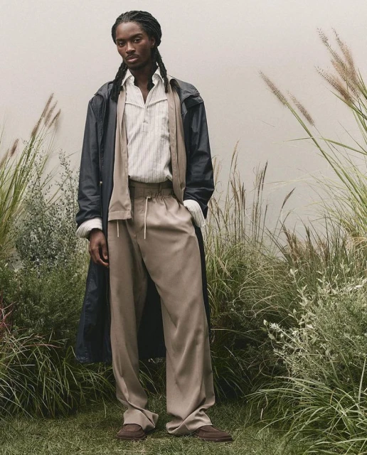 A stylish adult man in a button-down shirt, trench coat, and slacks poses in an outdoor setting with tall grasses and neutral backdrop.