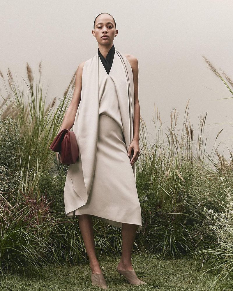 Elegant woman in a sleeveless cream-colored dress holding a clutch poses in tall grass with a neutral background, showcasing fashion.