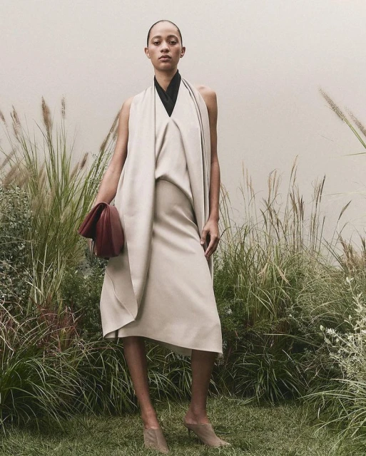 Elegant woman in a sleeveless cream-colored dress holding a clutch poses in tall grass with a neutral background, showcasing fashion.