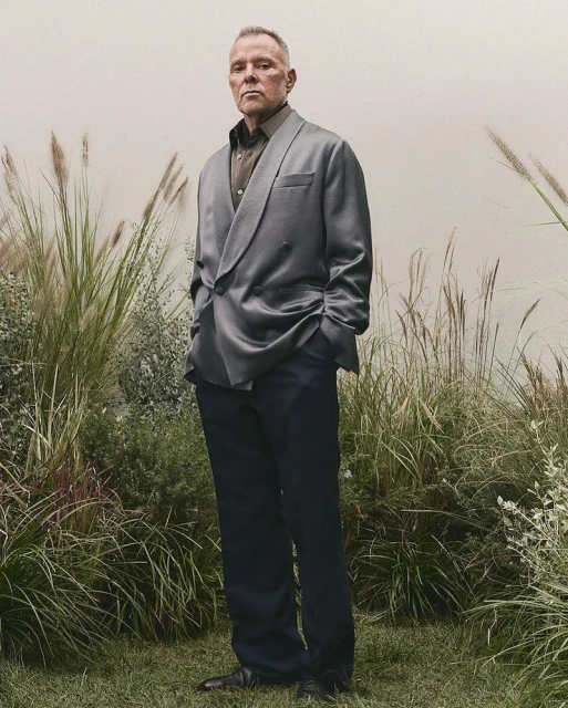 Full-length portrait of a grey-haired adult man in a double-breasted grey jacket and dark pants, standing in a grassy outdoor setting.