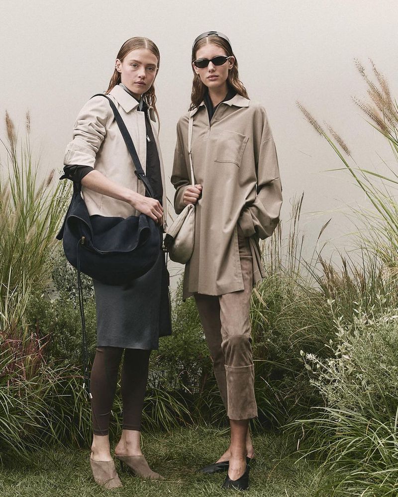 Two women model earth-tone fashions on a grassy field with tall grasses, one in a jacket and the other in an oversized shirt.
