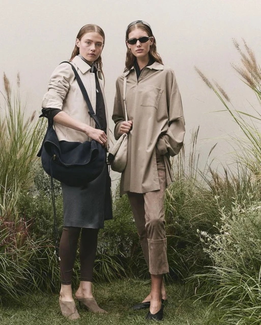 Two women model earth-tone fashions on a grassy field with tall grasses, one in a jacket and the other in an oversized shirt.