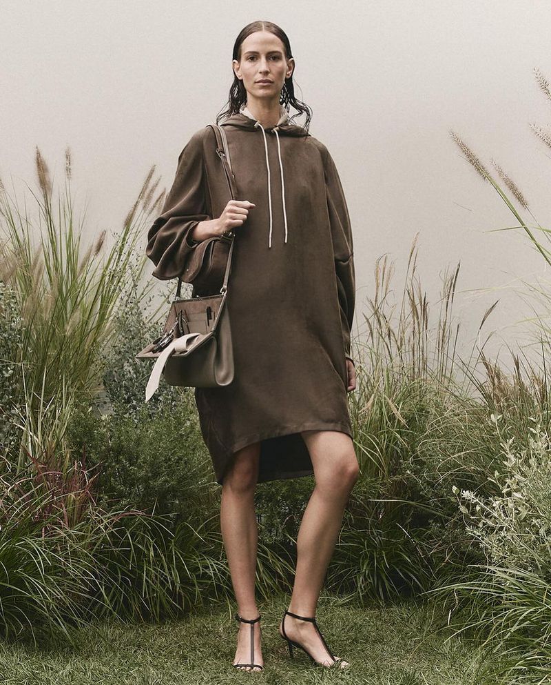 An adult woman stands in an outdoor setting, modeling a brown hoodie dress, a handbag, and black high-heel sandals for a fashion shoot.