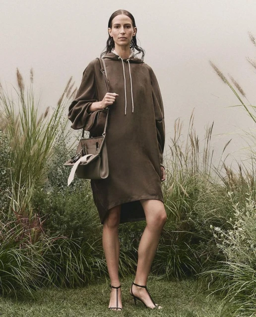An adult woman stands in an outdoor setting, modeling a brown hoodie dress, a handbag, and black high-heel sandals for a fashion shoot.
