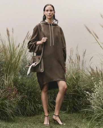 An adult woman stands in an outdoor setting, modeling a brown hoodie dress, a handbag, and black high-heel sandals for a fashion shoot.