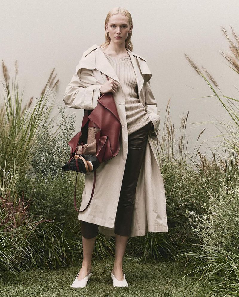A fair-skinned fashion model poses in a trench coat with a large handbag, set against a natural, grassy outdoor backdrop with soft lighting.