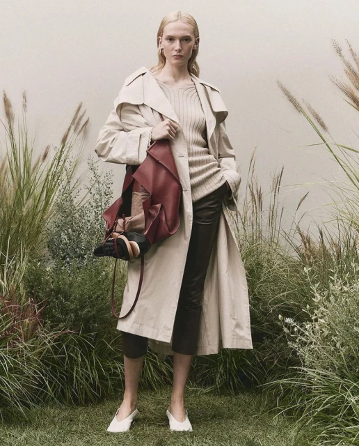 A fair-skinned fashion model poses in a trench coat with a large handbag, set against a natural, grassy outdoor backdrop with soft lighting.