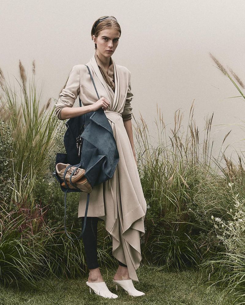 A woman poses in a neutral-toned coat with a bag and shoes against a backdrop of tall grass, illuminated with soft, natural light.