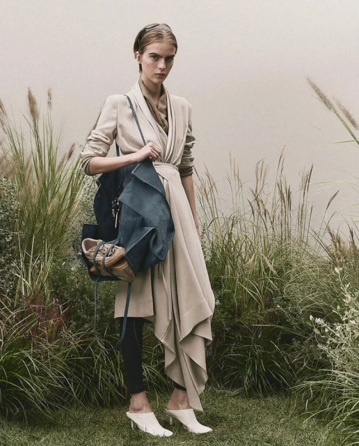 A woman poses in a neutral-toned coat with a bag and shoes against a backdrop of tall grass, illuminated with soft, natural light.