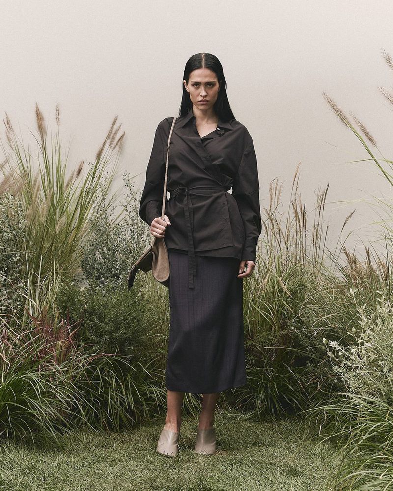 A brunette woman in a dark wrap shirt and skirt stands in a lush outdoor setting with tall grass and neutral background.