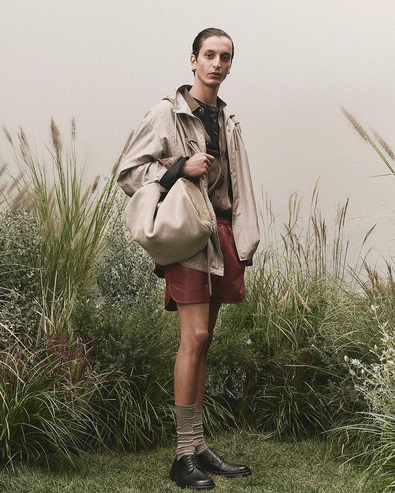 A fashionable adult male poses outdoors in a tan jacket, red shorts, socks, and black shoes, carrying a large tan handbag.