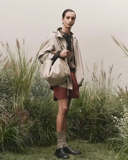 A fashionable adult male poses outdoors in a tan jacket, red shorts, socks, and black shoes, carrying a large tan handbag.