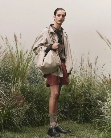 A fashionable adult male poses outdoors in a tan jacket, red shorts, socks, and black shoes, carrying a large tan handbag.