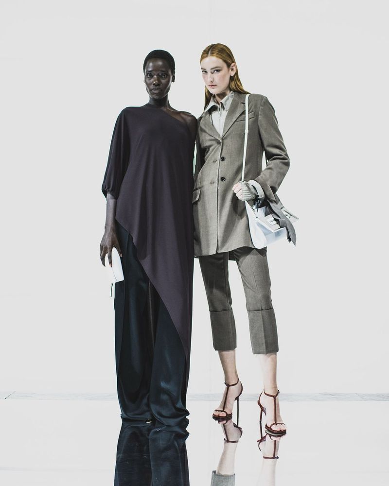 Two fashion models posing against a white background, wearing a dark draped top and a tailored grey suit jacket.