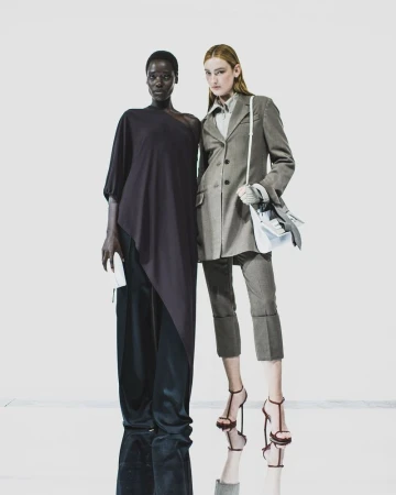 Two fashion models posing against a white background, wearing a dark draped top and a tailored grey suit jacket.