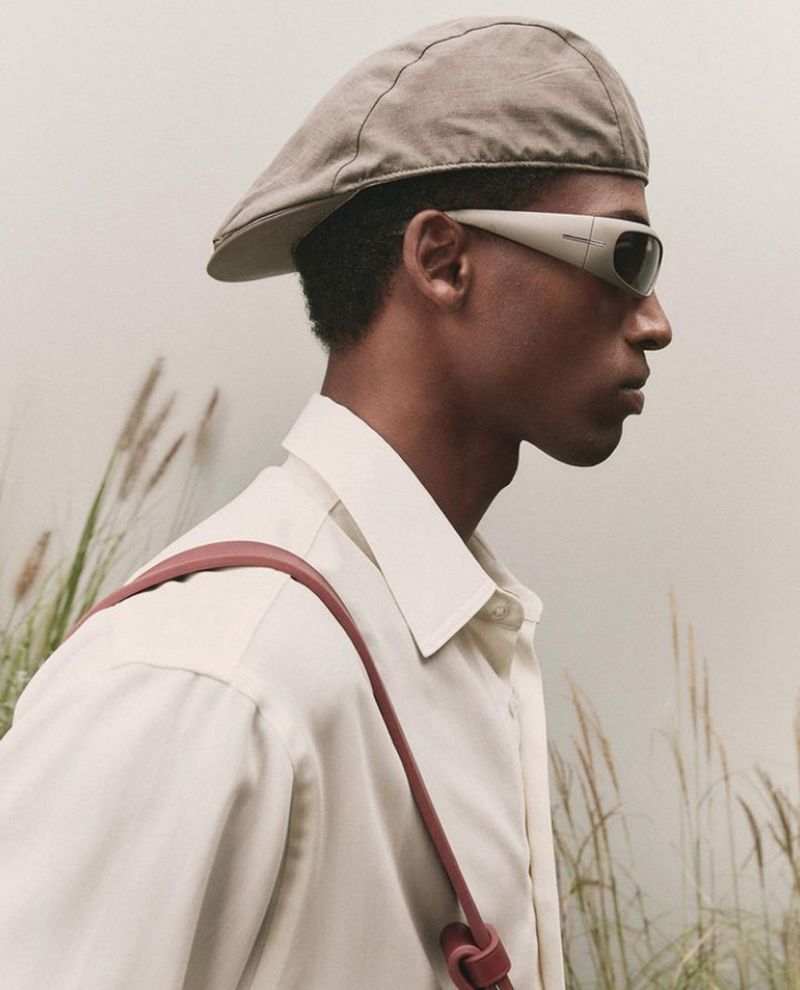 Person wearing sunglasses and a cap outdoors in a field with tall grass. The model is wearing a beige shirt and red shoulder bag.