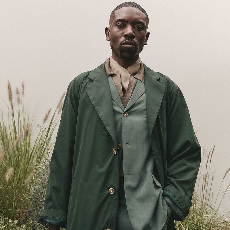 Adult male in a three-piece suit with a trench coat poses in front of tall green and brown grasses with a light grey backdrop.