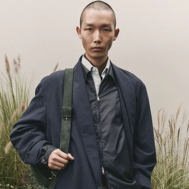 Adult male in a navy overcoat with a green bag, standing outdoors with a neutral background and tall golden grasses visible at the bottom.