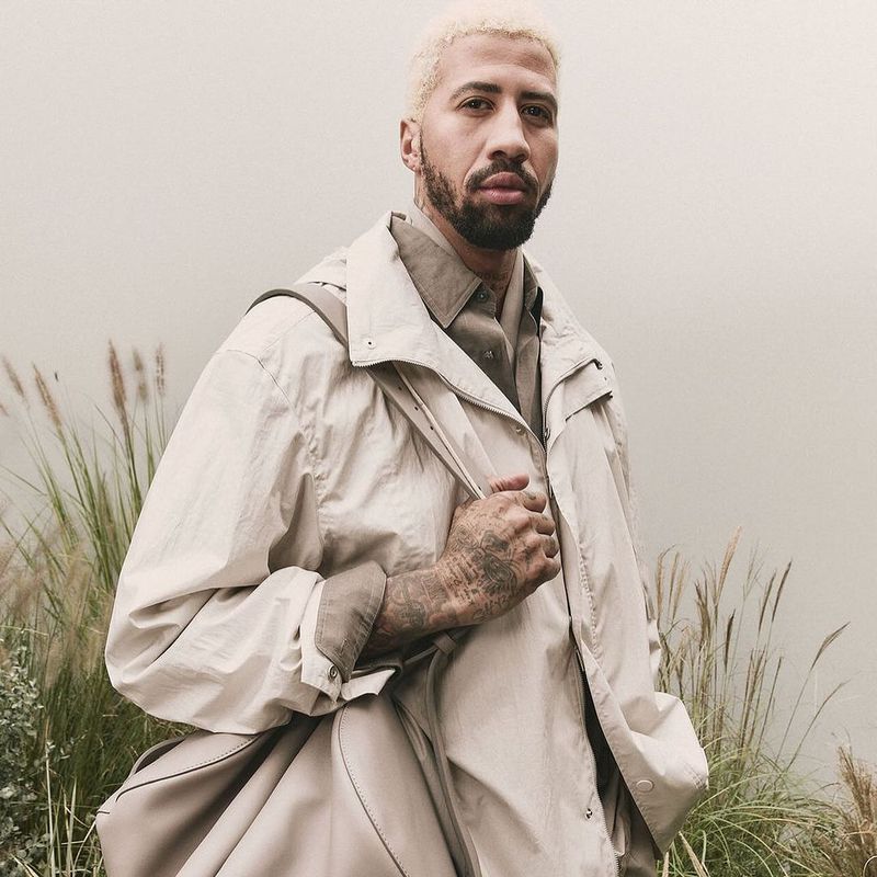 Adult male with bleached hair poses outdoors in a tan collared shirt and jacket while holding a tan leather bag over his shoulder.