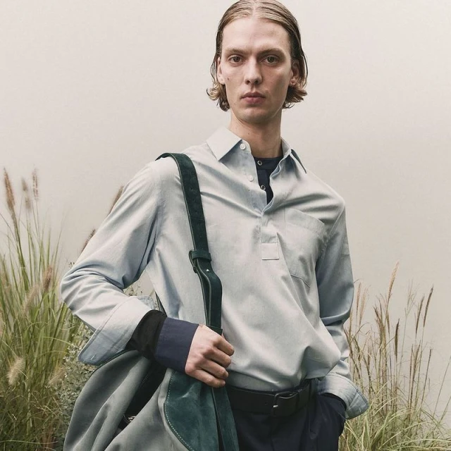 Fashionable adult male in a light blue button-down shirt with a dark green bag slung over his shoulder poses outdoors.
