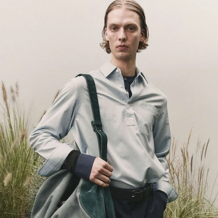 Fashionable adult male in a light blue button-down shirt with a dark green bag slung over his shoulder poses outdoors.