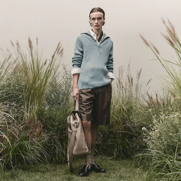A male model wearing a light blue ribbed sweater and dark shorts holds a beige tote bag amidst tall grass and plants.