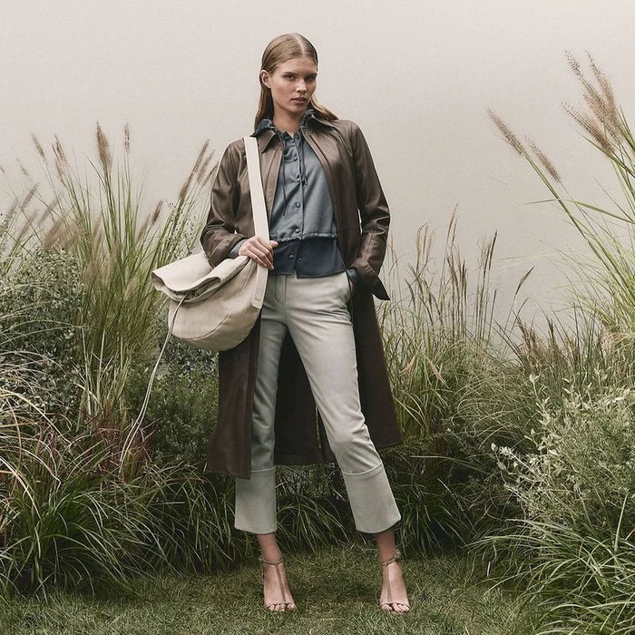 Model standing outdoors wearing a brown leather coat over a grey blouse and beige trousers, holding a cream shoulder bag.