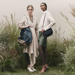 Two models standing among tall grass in neutral-toned apparel, holding dark handbags for Boss S/S 25 campaign.