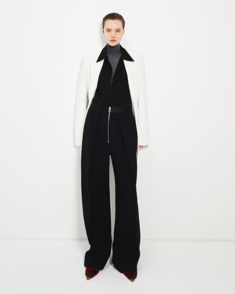 Full-length studio shot of a female fashion model wearing a white jacket, gray turtleneck, and black wide-legged pants against a white backdrop.