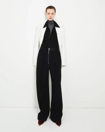 Full-length studio shot of a female fashion model wearing a white jacket, gray turtleneck, and black wide-legged pants against a white backdrop.