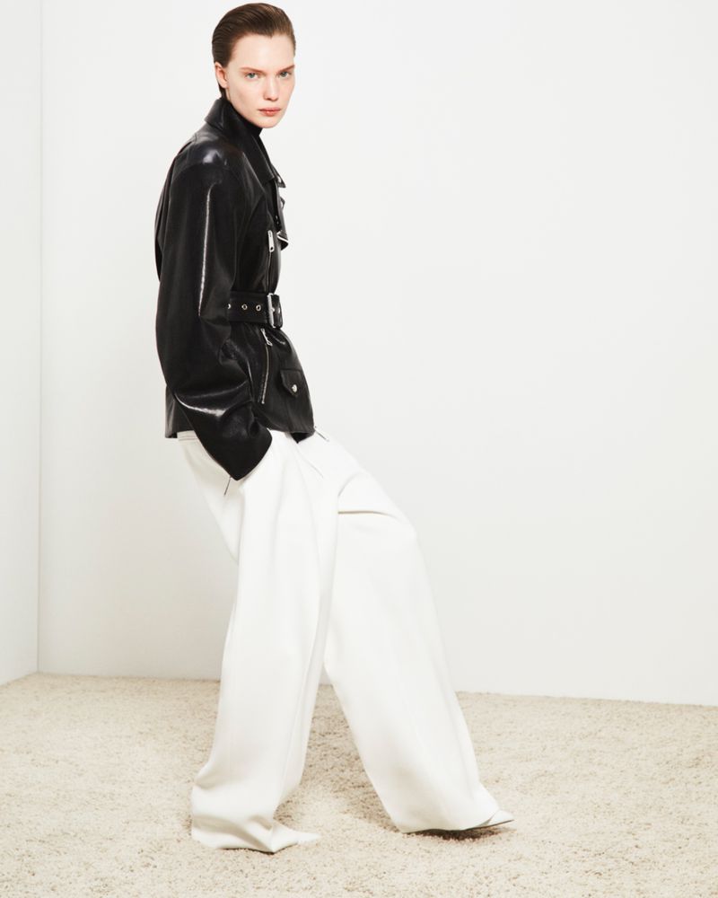 Fashion shot of a model wearing a shiny black leather jacket and wide white trousers against a white backdrop in a studio environment.