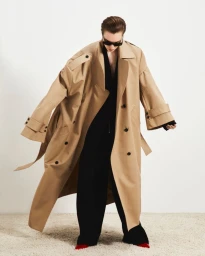 Model wearing dark sunglasses and an oversized tan trench coat over black garments against a white studio background.