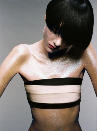 Woman with short black hair and glitter makeup wearing a strapless bandeau top in a studio setting, looking down with neutral expression.