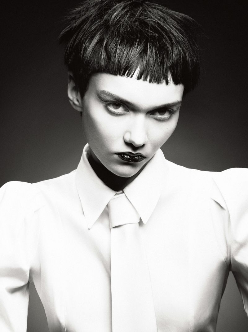 Monochrome portrait of a young adult woman with short dark hair, dramatic makeup, and a white collared shirt with a tie against a grey backdrop.
