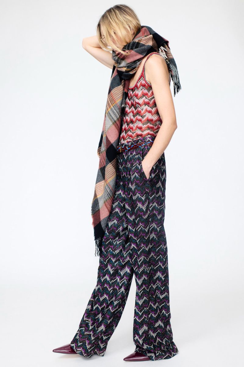 Fashionable woman in patterned top and pants poses with a large plaid scarf against a white background, hands near head, looking away.