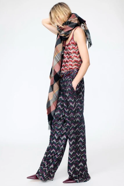 Fashionable woman in patterned top and pants poses with a large plaid scarf against a white background, hands near head, looking away.