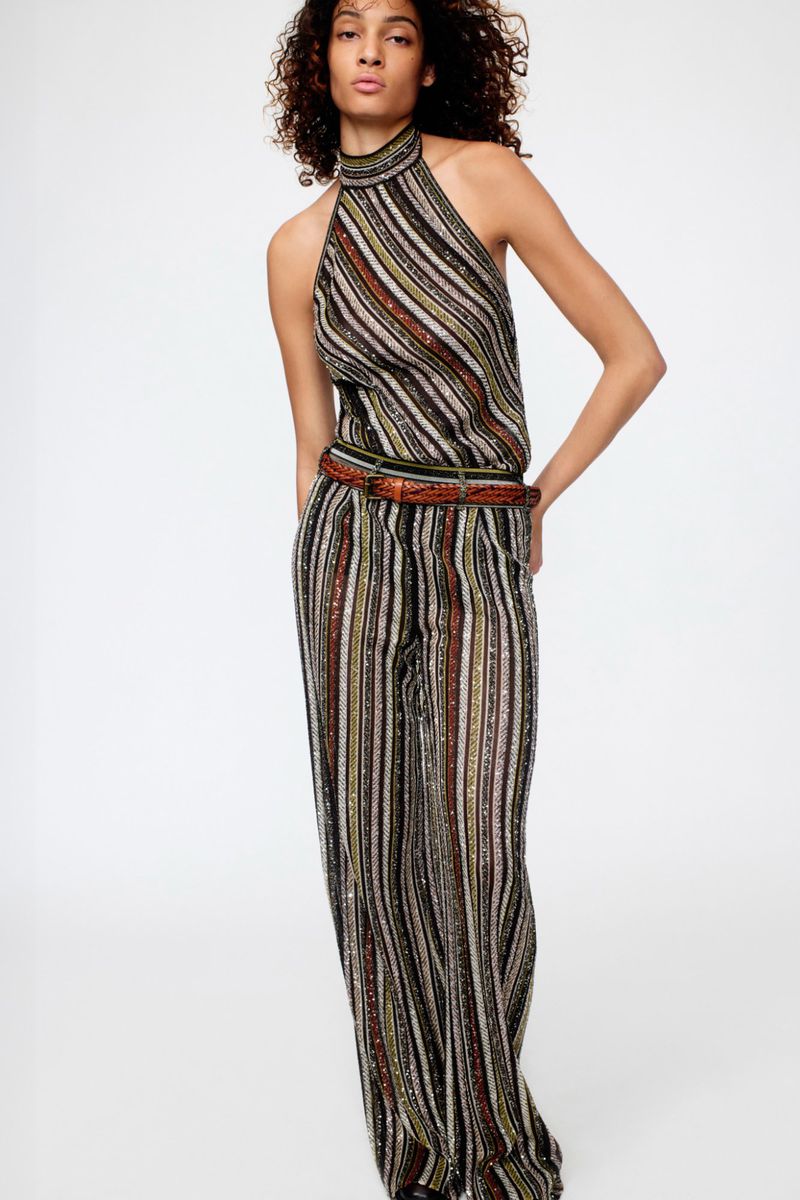 Fashion shot of an adult female wearing a striped halter jumpsuit with a brown belt, posing in front of a plain background in studio lighting.