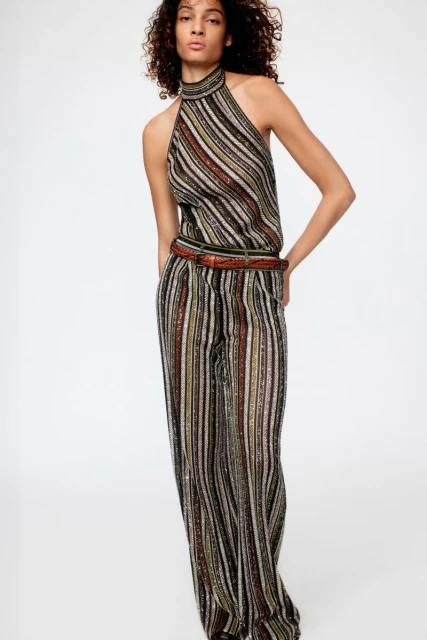 Fashion shot of an adult female wearing a striped halter jumpsuit with a brown belt, posing in front of a plain background in studio lighting.