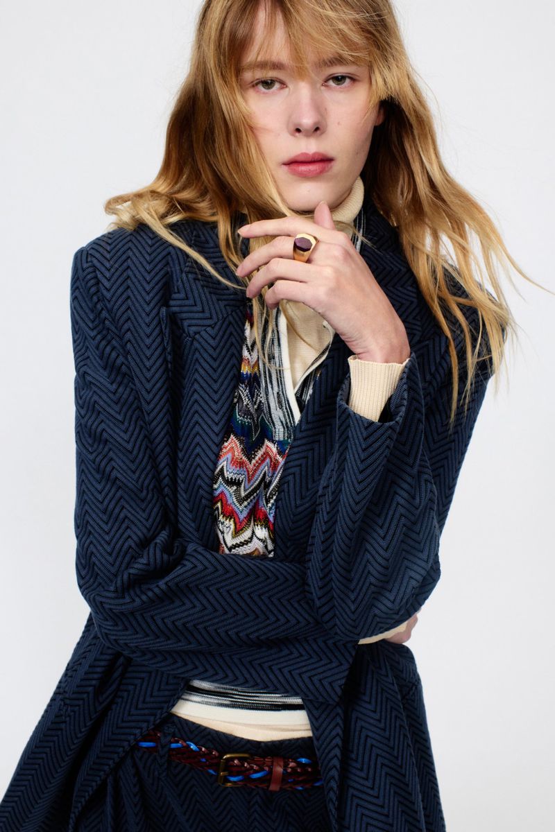 A fashionable adult female with wavy blonde hair wearing a blue patterned coat and ring poses indoors against a clean, white background.