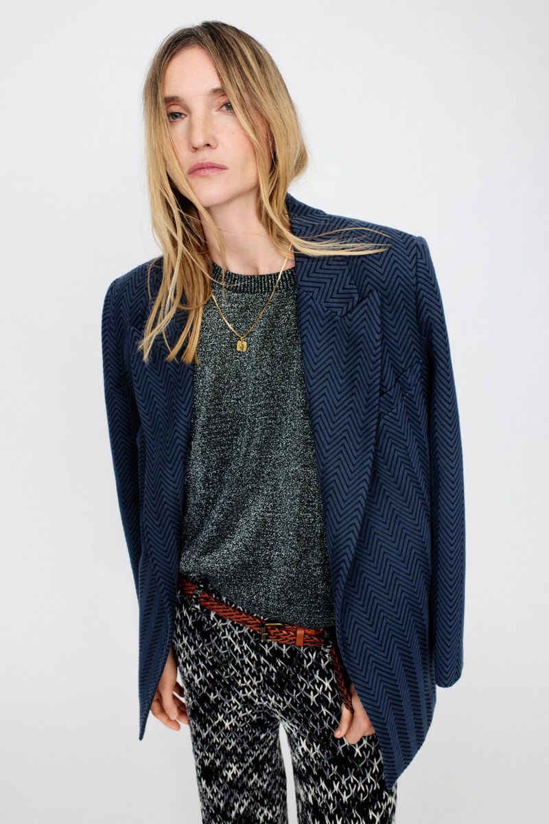 Model wearing a structured navy blazer over a dark sweater and patterned trousers against a white studio background.