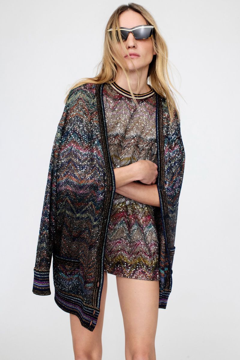 Adult woman poses indoors wearing colorful, glittering knitwear cardigan and matching dress, paired with futuristic sunglasses in a studio shot.