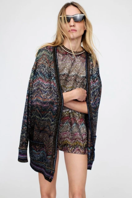 Adult woman poses indoors wearing colorful, glittering knitwear cardigan and matching dress, paired with futuristic sunglasses in a studio shot.