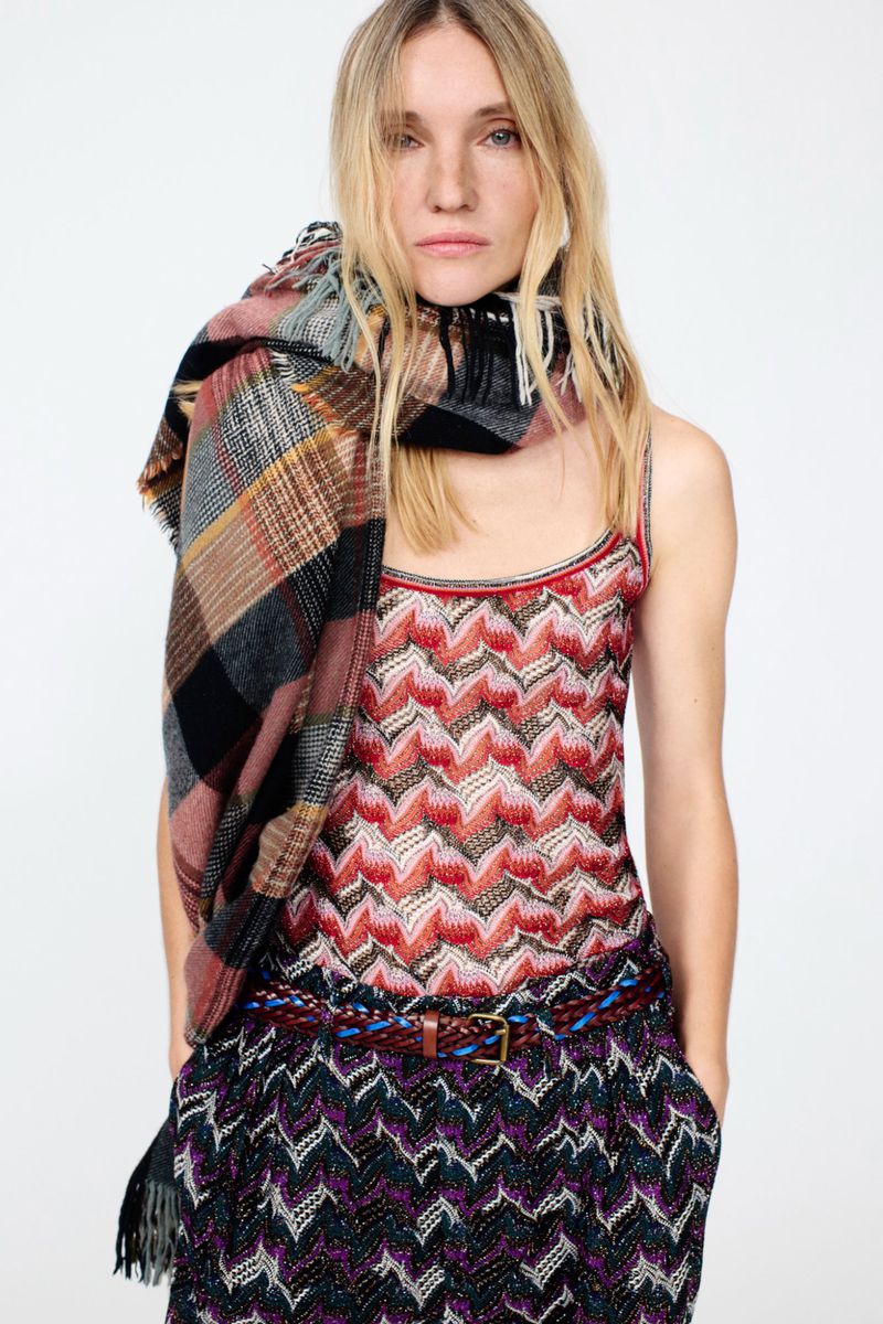 A fashionable woman is wearing a patterned top and pants with a plaid scarf. She has blonde hair and a neutral facial expression.